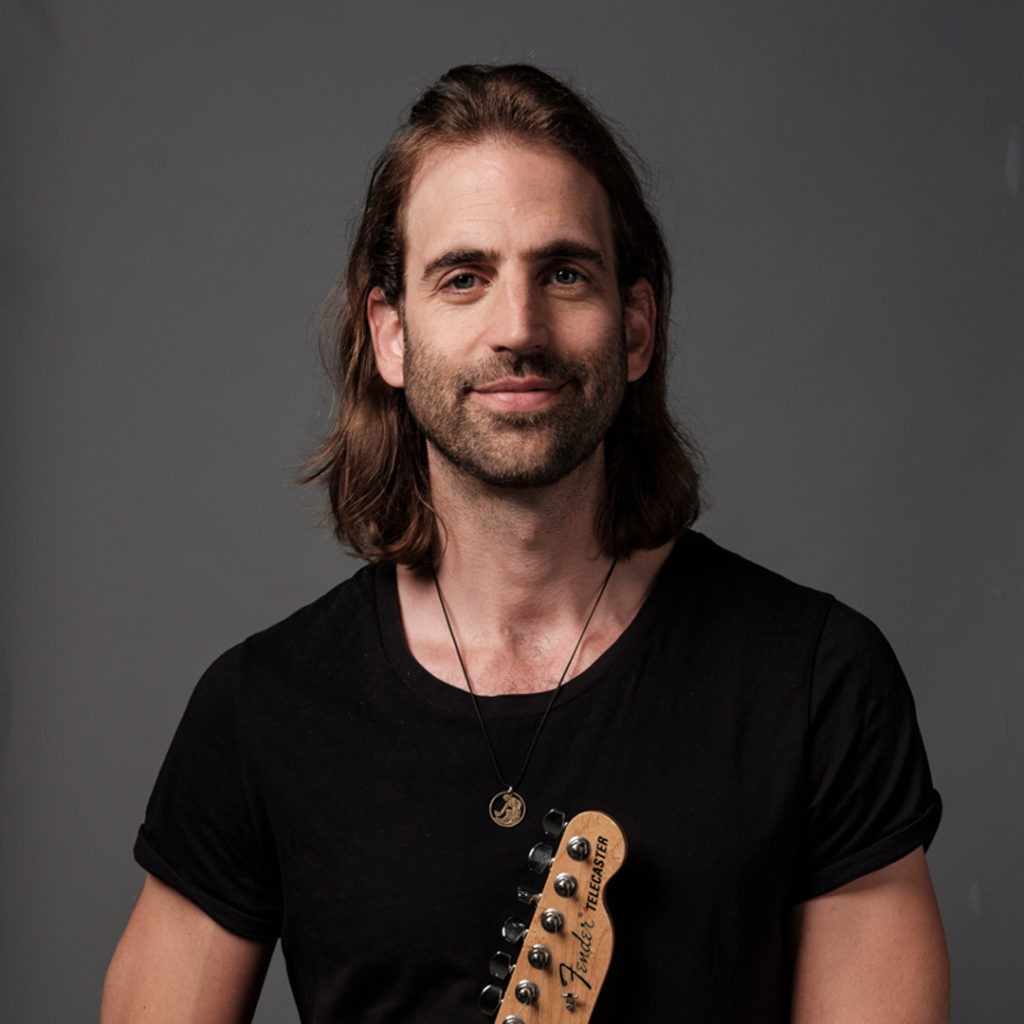 Jessy Hay is gitarist bij Marmoucha Orchestra. Jessy staat op deze foto voor een grijze muur met een gitaar in zijn handen.