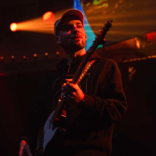 Johnny Biner is gitarist bij Marmoucha Orchestra. Johnny staat op de foto met rode lichten op de achtergrond en met een elektrisch gitaar in zijn handen.