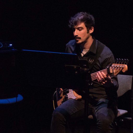 José Santibañez is bassist en componist bij Marmoucha Orchestra. José zit op deze foto op een stoel met een zwarte achtergrond achter zich en spelend op de gitaar.
