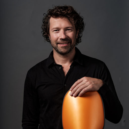 Lucas Stam is cellist bij Marmoucha Orchestra. Lucas staat op de foto voor een grijze muur met een cello in zijn handen.