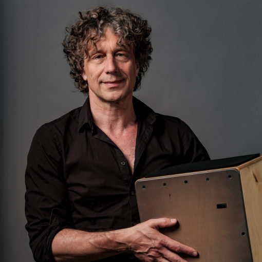 Udo Demandt is crossover-percussionist & drummer in Marmoucha orchestra. Udo staat op deze foto voor een grijze muur met een instrument in zijn handen.