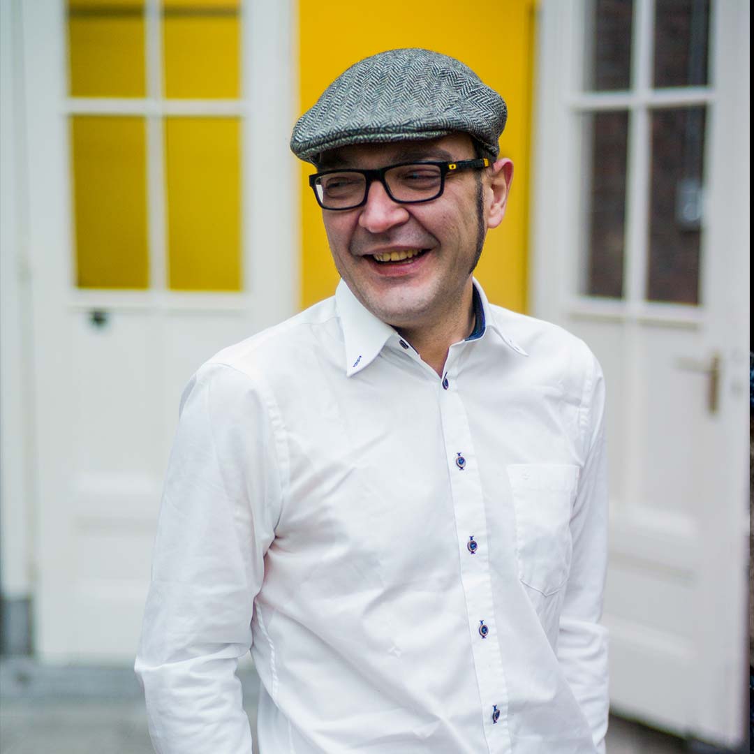 Daniele Cappucci is contrabassist bij Marmoucha Orchestra. Daniele staat op deze foto voor witte deuren. Daniele heeft een witte blouse aan en een grijs petje op.